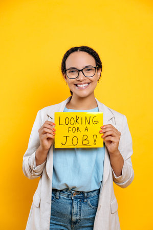 Vertical photo of positive successful smart mixed race young woman, creative specialist looking for a job, stands on isolated orange background, shows a sign looking for a job, looks at camera, smileの写真素材