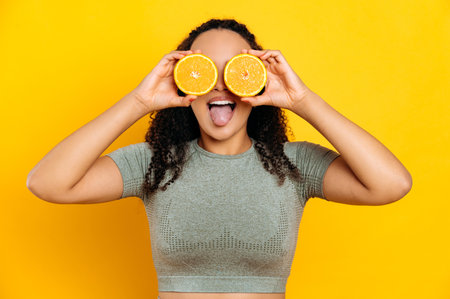 Joyful lovely brazilian or latino young curly haired woman, in sport outfits, standing on isolated orange background, holds two halves orange in her hands near eyes, shows tongue, smilingの写真素材