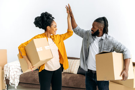 Joyful happy married african american family couple, holding boxes with things for the house, stand...