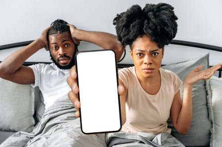 Puzzled upset african american couple, spouses, sit in a cozy bed in the bedroom, woman shows smartphone with blank mock-up screen, space for advertising, presentation, look at camera at a lossの写真素材