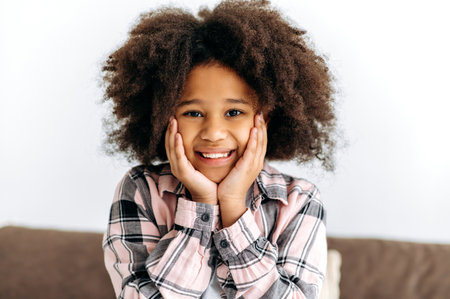 Photo of a cute beautiful African American preschool girl, with curly hair, in a basic clothes, sits on the sofa, looks to the camera, smiles friendly. Happy childhoodの写真素材