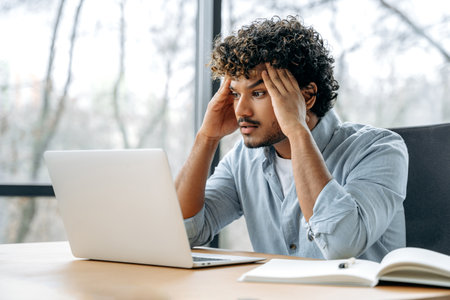 Tired exhausted overworked indian office worker, creative manager, sitting at workplace holding his...