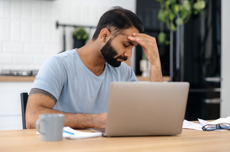Overworked fatigue upset indian or arabian man, manager or freelancer, sit at a desk in the kitchen with laptop, with eyes closed, in need of rest in a stressful situation, have headache, migraine,の写真素材