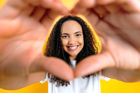 Gorgeous positive brazilian or hispanic curly haired woman, demonstrates with hands the symbol of heart, love, looks through it at the camera, smiles friendly, stands on isolated yellow backgroundの写真素材