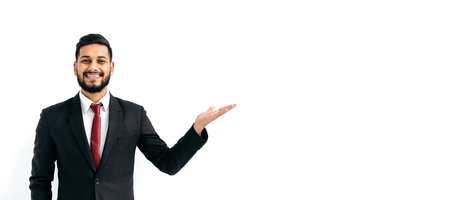 Copy-space for your presentation. Panoramic photo of positive elegant arabian or indian business man, stand on isolated white background, looks at camera, pointing hand to empty space, smiles happilyの写真素材