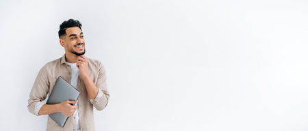 Panoramic photo of handsome arabian or indian guy in stylish casual wear, freelancer or student, holding a laptop, stand on white isolated background, looking away, smiling happily. Copy-space conceptの写真素材