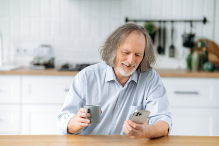 Satisfied senior caucasian grey haired man with beard, sits at home in the kitchen at the table,...