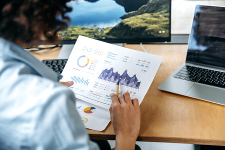 Back view of successful african american female stock investor, broker, financial expert, sit at work desk, uses computer and charts, analyzes risks and prospects, rise or fall of cryptocurrencyの写真素材