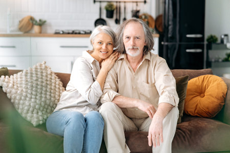 Happy together. Modern pensioners. Middle aged caucasian married couple, grey haired spouses spend time together in living room, wife and husband sit on a sofa, hugging, looking at camera, smileの写真素材