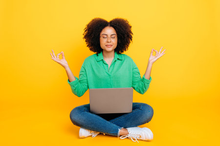 Full-length photo of calm relaxed african american or brazilian curly woman, stylishly dressed, sits on a yellow background with a laptop, meditate with eyes closed, take care of mental health, smilesの写真素材