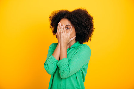 Scary, afraid. Terrified hispanic or brazilian curly haired woman dressed in casual shirt, peeks through fingers, covers face with hands, experiencing scare, shock, stand on isolated yellow backgroundの写真素材