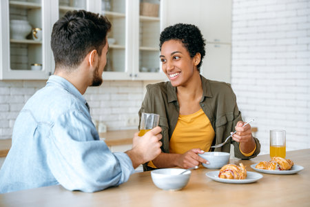 Happy young family couple, caucasian man and african american woman, having breakfast at cozy home kitchen with healthy porridge, happy to spend time together at weekend morning, communicating, smileの写真素材