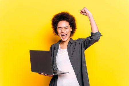 Joyful african american or brazilian young woman with short curly hair, holding laptop, gesturing with hands, rejoices at victory, success, profit, stand on isolated yellow background, smilingの写真素材