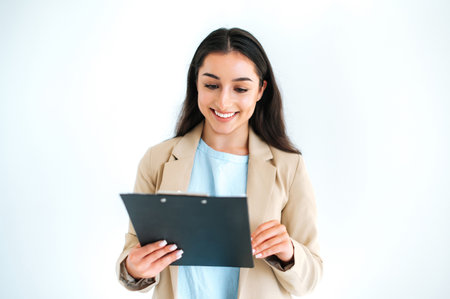 Happy beautiful brunette indian or arabian woman, hiring manager, financial consultant stands on isolated white background, holding folder with documents or resume, looking at it, smiling friendlyの写真素材