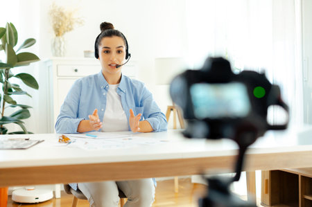 Confident young woman, blogger, influencer sits at a desk, looking at camera, recording education...