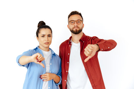 Displeased arabian man and caucasian woman showing thumbs down, expressing disapproval and disappointment, dressed casually, stand on isolated white background, looking at cameraの写真素材