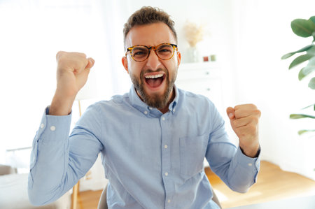 Joyful caucasian or arabian man with glasses in a blue shirt, raising his fists in excitement,...