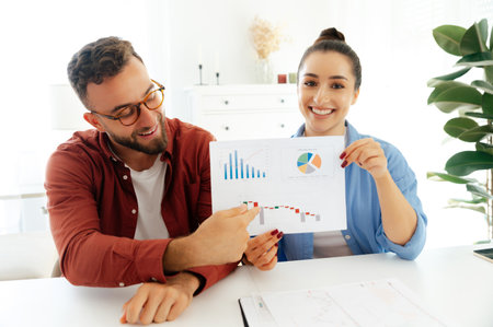 Two friendly confident coworkers in stylish clothing, are sitting at a desk in an office, showing a financial chart to camera during video call, online financial lesson, recording financial lectureの写真素材