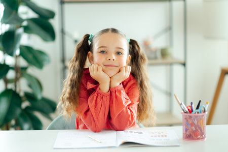 Cute blue-eyed schoolgirl in a red blouse rests her chin on hands while sitting at a desk with a workbook at home, looking calm and thoughtful, smiling at the cameraの写真素材