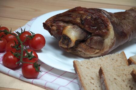 Close view photo of tasty ham hock or pork knuckle from a pig with knife bread and fresh cherry tomatoes on wooden desk. Smoked dried meat food.の写真素材