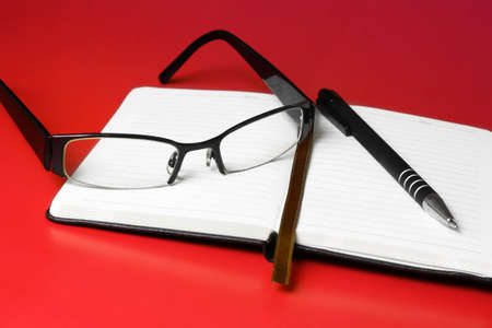 Close up of reading glasses and pen on top of open diary isolated on red backgroundの写真素材
