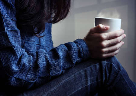Young woman sitting and waiting with cup of hot drink in her handsの写真素材