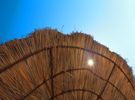 edge of beach umbrella from dry grass with translucent sun on blue sky background