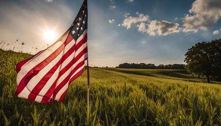 A vibrant American flag waves proudly in a field of lush green grass, bathed in the warm glow of a setting sun. The sky is a beautiful blend of blue and golden hues, with fluffy white clouds floating across its expansion.の写真素材