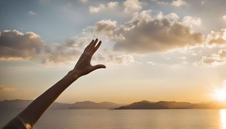 A single hand stretches towards the setting sun over a tranquil body of water, creating a powerful silhouette against the sky.の写真素材