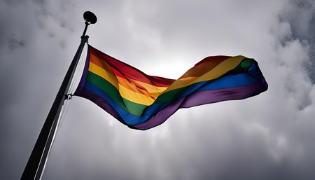A vibrant rainbow pride flag waves proudly in the wind against a cloudy sky, symbolizing diversity,...
