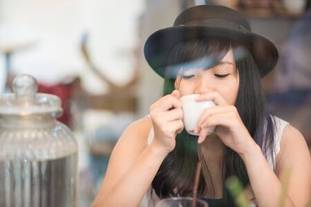 Young asian woman drinking coffee through the mirror in a trendy vintage cafeの写真素材