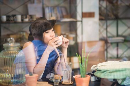 Hipster asian woman drinking coffee in cafe through mirrorの写真素材