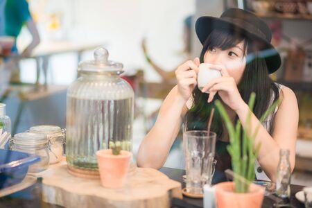 Hipster asian woman drinking coffee in cafe through mirrorの写真素材
