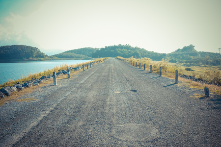 Rural road near river of dam under sunny skyの写真素材