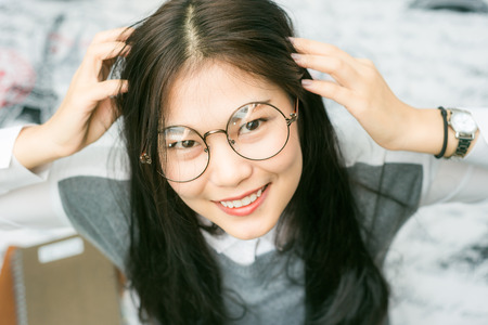 Beautiful asian young curly woman with glasses sitting on the bed for reading bookの写真素材