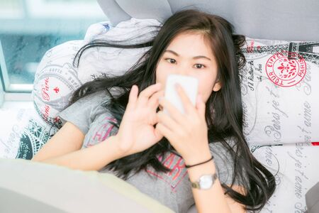 Beautiful black asian woman using on her phone laying in her bedroom, smiling asian womenの写真素材