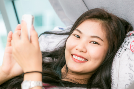 Beautiful black asian woman using on her phone laying in her bedroom, smiling asian womenの写真素材