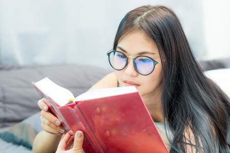 Attractive nerd student woman on bed reading book at cozy room