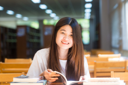Happy asian student  young woman thinking with book in library sitting on wood tableの写真素材
