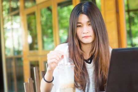Happy smiling woman working with laptop and drinking cold latte coffee on wood table at coffee shopのeditorial素材