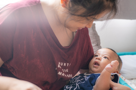 Asian women holding smiling infant boy in bedroom morning lightの写真素材