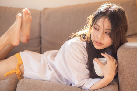 Attractive beautiful asian women with coffee cup lying on sofa morning sun lightの写真素材