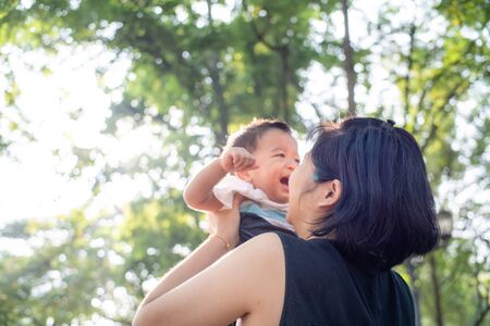 Mom lifting hold infant baby boy in city park against sun light morning