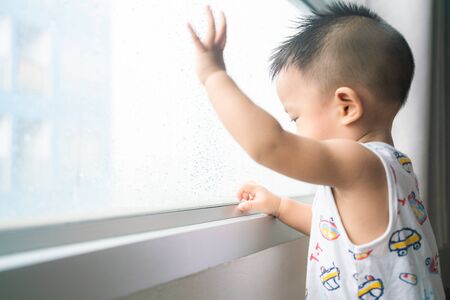 Happy baby boy stand near window looking to rainy outside from condominiumの写真素材