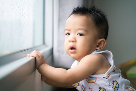 Happy baby boy stand near window looking to rainy outside from condominiumの写真素材