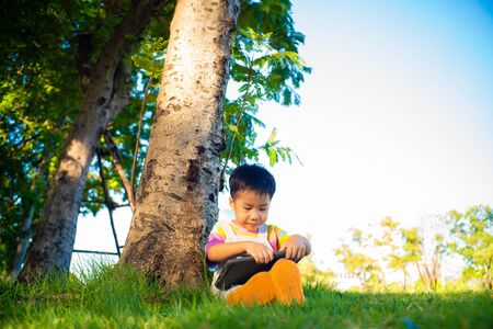 Adorable asian boy sweet casual sitting on green grass use tablet computer learnning nature tree in...