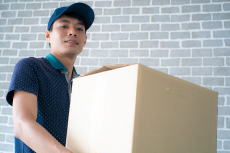 Happy young delivery man in blue cap standing with parcel post box in houseの写真素材