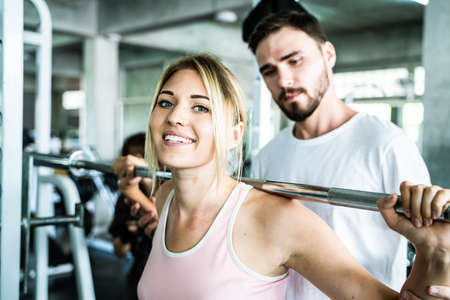 Cheerful young woman wearing pink sports bra while doing chin-up exercise with trainer man in fitness gymの写真素材