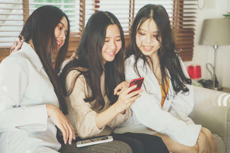 Beautiful asian women use smartphone social media together sitting on sofa in cozy roomの写真素材