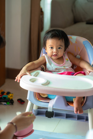 Mother Feeding Baby Girl with a Spoon Giving Food to adorable Child Sitting on chair at Home. Baby...